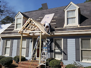A crew member kneeling on a roof setting wooden trusses for a new porch roof, with two ladders below